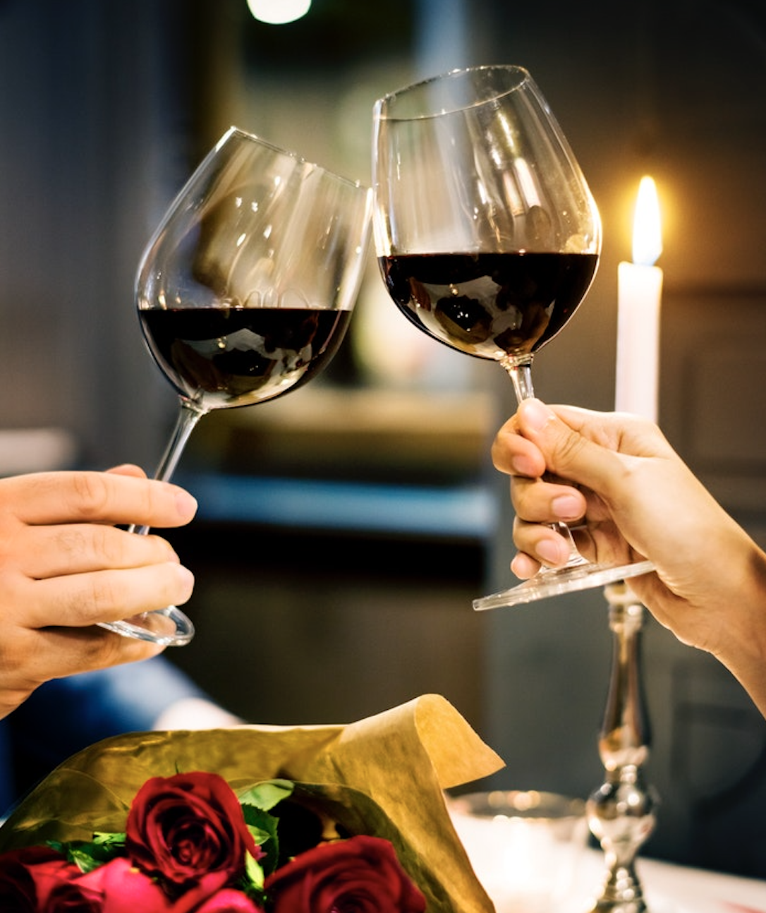 Two glasses of red wine toasting by candlelight next to a bouquet of roses